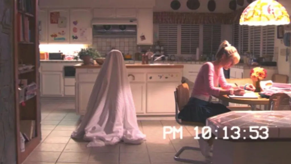 A teenager sits at a kitchen table doing homework while a child covered in a bedsheet is behind her